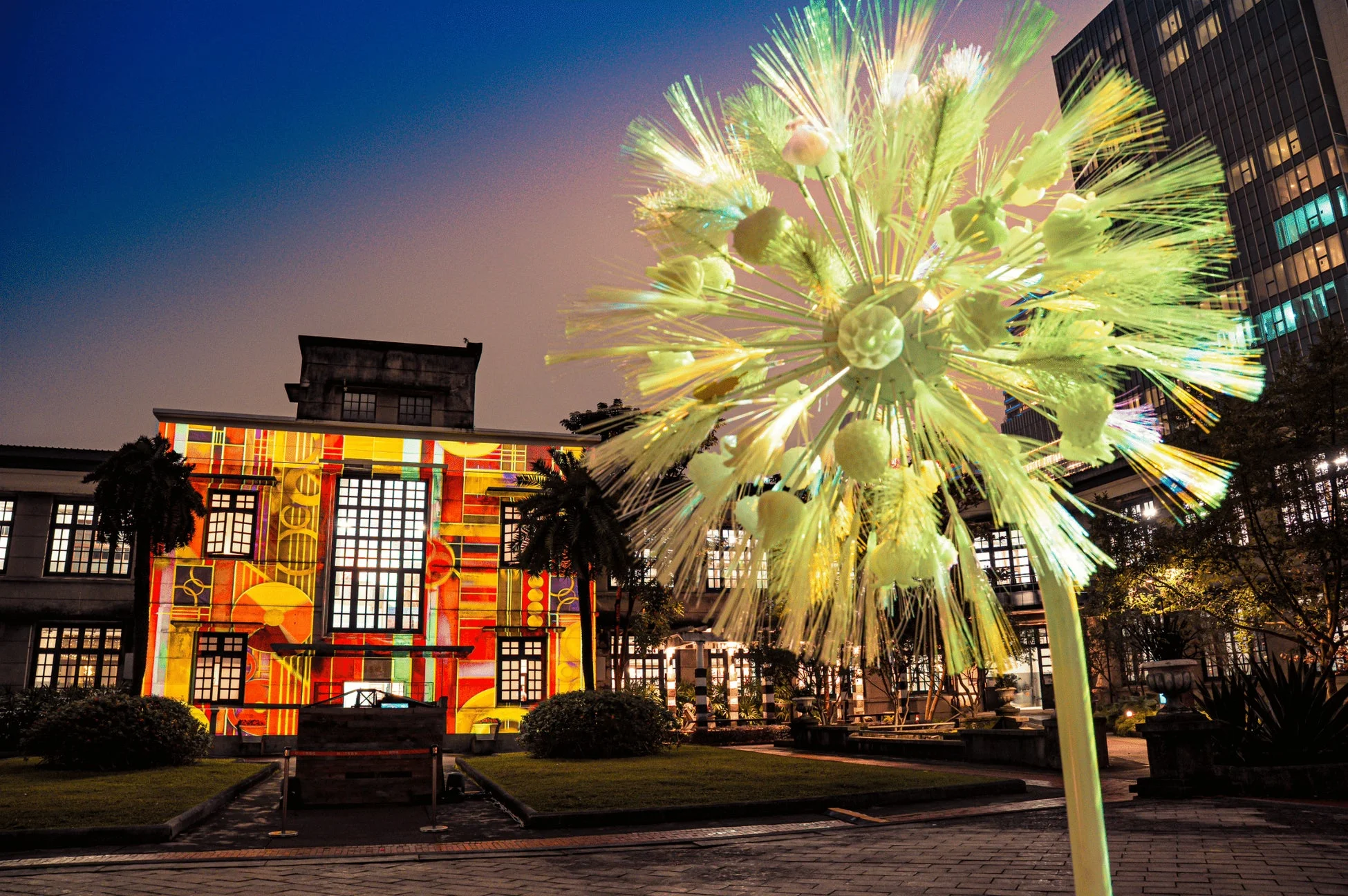 夜の都市公園で、カラフルなプロジェクションマッピングが施された建物と、タンポポのような形状の光るアートオブジェが印象的な一枚