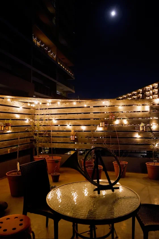 月が輝く夜空の下、ストリングライトで飾られたバルコニーまたはテラスの夜景です。温かい光が木製の壁と植木鉢を照らし、ガラスのテーブルと椅子がリラックスした雰囲気を醸し出しています。