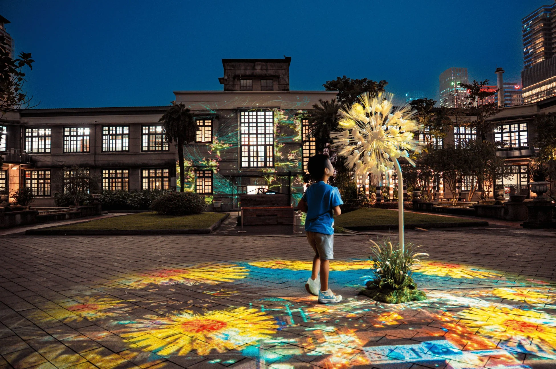夜の広場で、子供が光るタンポポのオブジェと建物に投影されたカラフルなプロジェクションマッピングを眺めている幻想的な光景