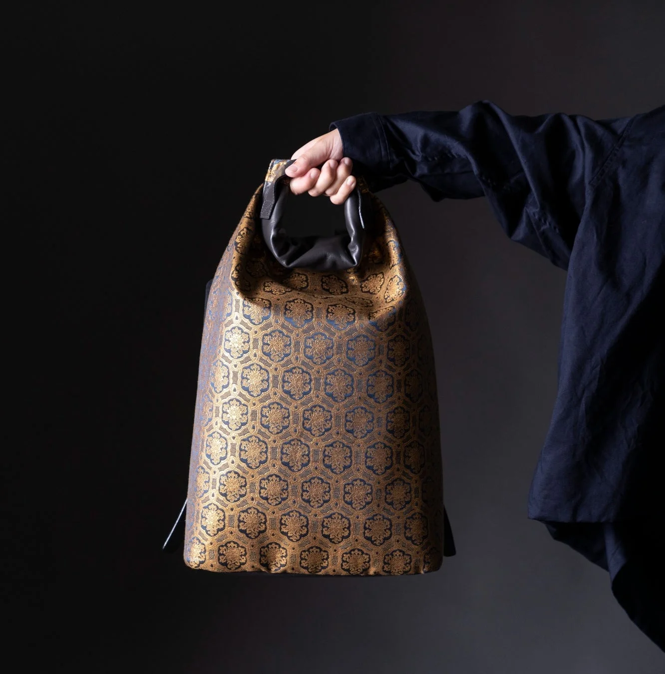 日本の伝統的な亀甲花菱のような幾何学模様が施された金色と紺色のバッグを、黒い服を着た人物が手で持っている様子
