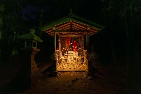 夜の暗闇に包まれた森の中に佇む小さな祠が、内部の灯りで明るく照らされている