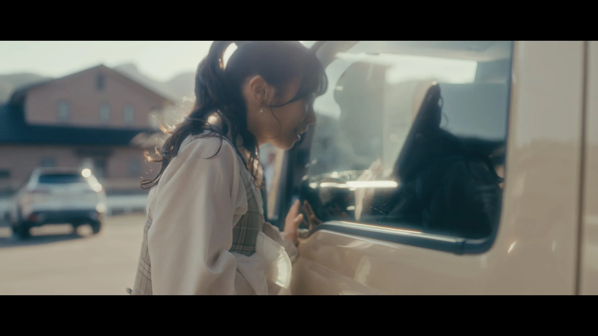 屋外で、一人の若い女性がベージュ色の車の窓を覗き込み、何かを話しているかのように笑顔を見せています。背景には建物と山が見え、柔らかな日差しが差し込む穏やかな情景です。