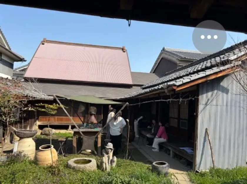 青空の下、伝統的な日本家屋の庭で人々が集まっている様子を捉えた画像です。庭には陶器や植物があり、縁側では数人が座ってくつろいでいます。古民家のような趣のある風景が広がっています。