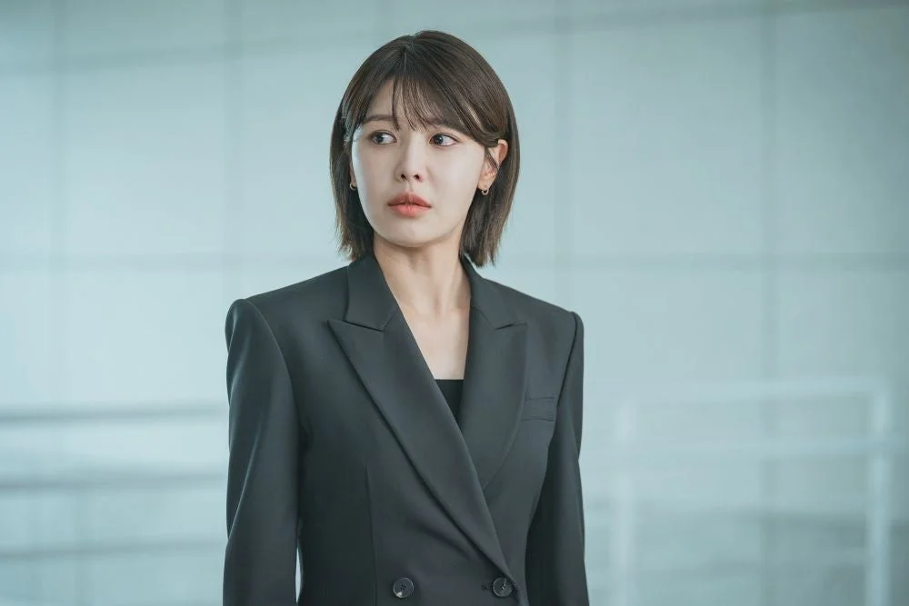 黒いダブルのブレザーを着たショートヘアの女性が、やや真剣な表情で横を見ているポートレートです。背景は明るい色の屋内空間で、ビジネスシーンを思わせる雰囲気です。