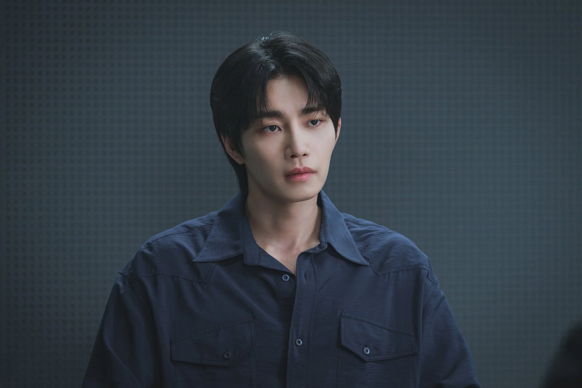 暗い青色のシャツを着た若いアジア人男性のポートレートです。黒髪で、やや斜め上を見つめるような無表情な顔つきをしています。背景はシンプルな暗い色の壁です。