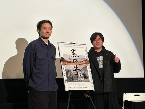 映画「黒の牛」のポスターを挟んで立つ2人の男性
