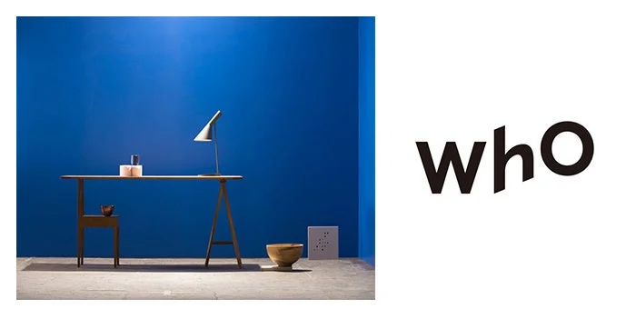 鮮やかな青い壁の部屋に、ミニマリストな木製デスクと白いデスクランプが配置され、右側には「who」というロゴが表示されている画像