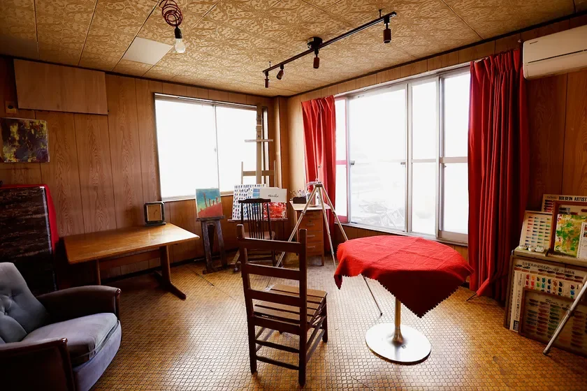 アトリエのような部屋で、イーゼルや画材、絵画が置かれ、創作活動が行われている様子を思わせる。木目調の壁やレトロな家具が特徴的で、窓から光が差し込んでいる。