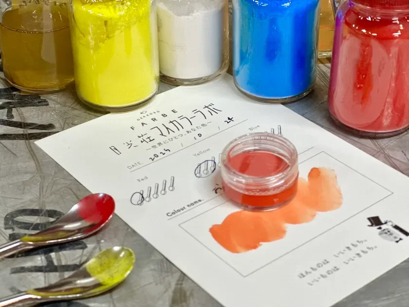 月光荘のカラーラボで、様々な顔料を使って自分だけのオリジナルカラーを作るワークショップの様子を捉えた写真です。カラフルな瓶に入った顔料や、作成中の赤色の色見本、日付が記された紙が写っており、色彩豊かな体験が伝わります。
