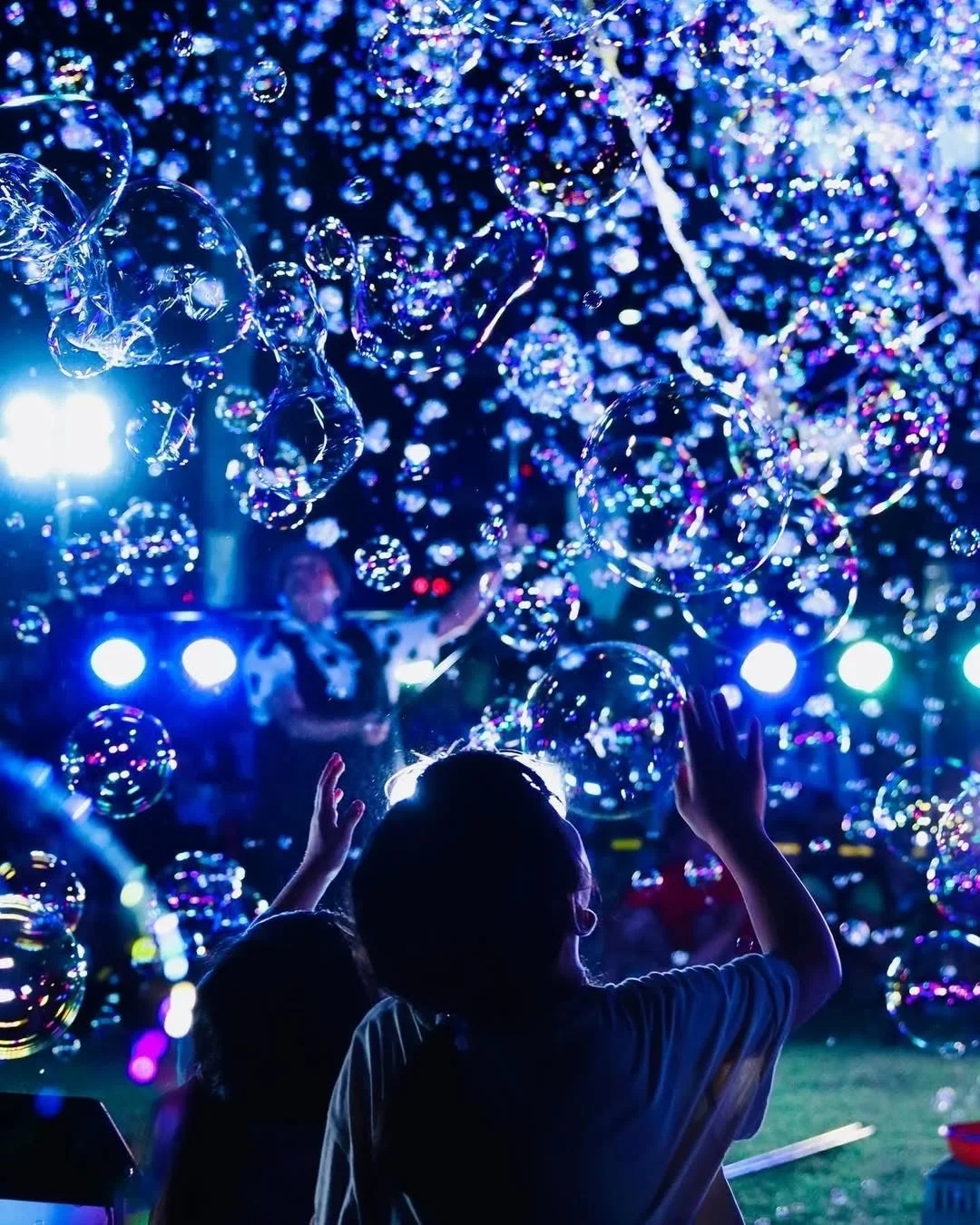 青い光に照らされた無数のシャボン玉が舞い上がる中で、人々が手を伸ばしてその美しさや楽しさを満喫している夜のイベントの様子です。