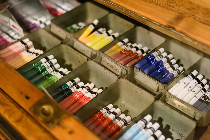 木製の引き出しに、色とりどりの絵の具チューブが金属製の仕切りに整理されて収納されています。緑、赤、青、黄、白など様々な色の画材が並び、絵を描くための道具が豊富に揃っている様子が伺えます。
