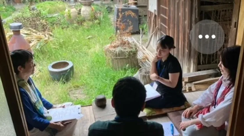 縁側のような場所で数人の男女が書類を手に話し合っている様子です。屋外の緑が見える、和やかな雰囲気の会議風景が捉えられています。