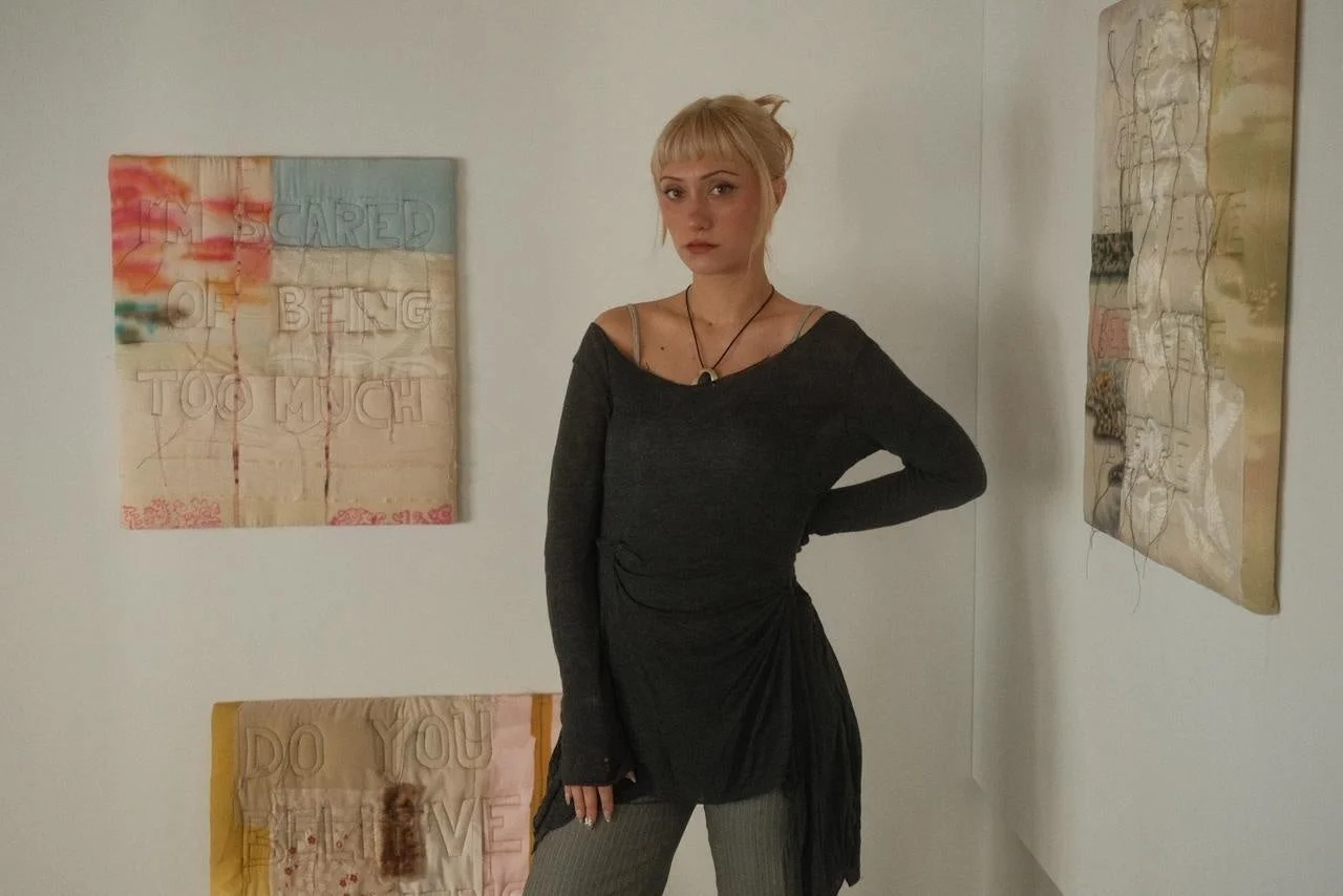 金髪の若い女性が白い壁の部屋でポーズをとっています。背後には「I'M SCARED OF BEING TOO MUCH」と書かれたアート作品などが飾られています。