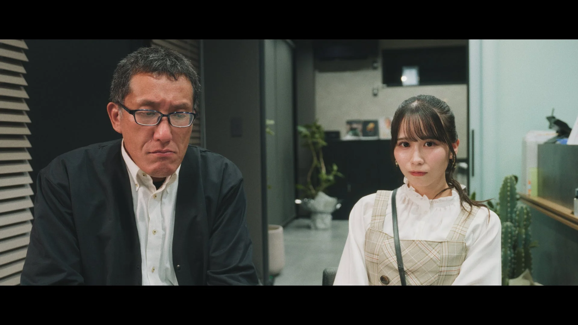 メガネをかけた中年の男性と若い女性が、真剣な表情でカメラを見つめています。室内で、打ち合わせのような状況に見えます。