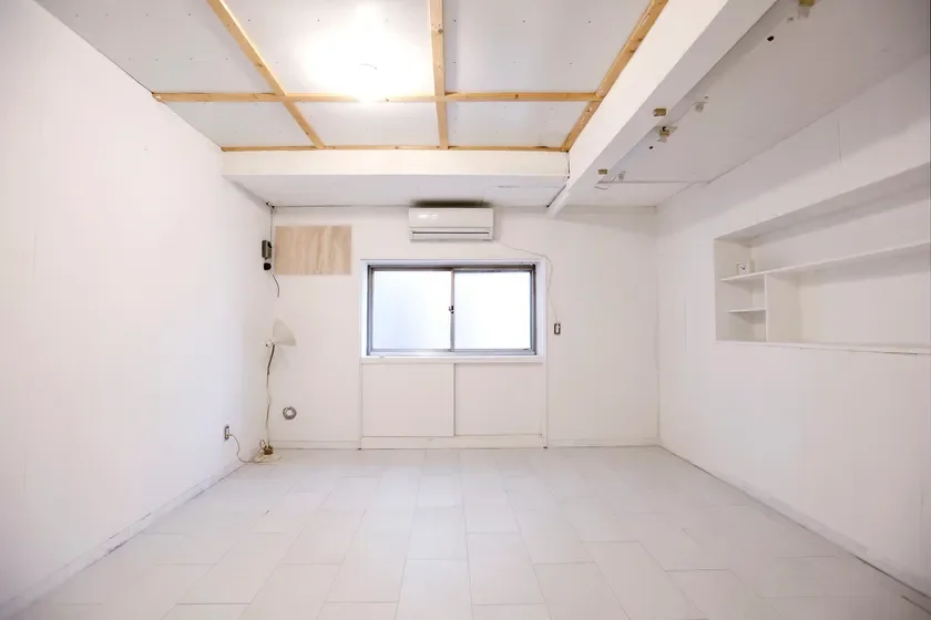 白い壁とライトグレーのタイル床が特徴の、広々とした空室です。窓の上にはエアコンが設置されており、右手の壁には備え付けの白い棚があります。天井の一部に木製の格子が見えるシンプルな内装で、様々な用途に活用できそうな空間です。