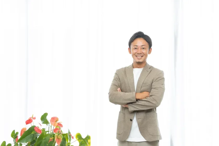 笑顔の男性が腕を組んで立っており、背景には白いカーテンとアンスリウムの鉢植えが見えます。