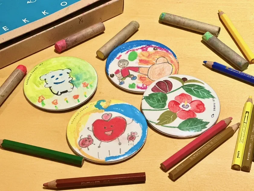 色鉛筆やクレヨンなどの画材と、子供が描いたような可愛らしい絵や花の絵が施された丸い作品が並べられています。手作り感のある温かいアートの風景です。