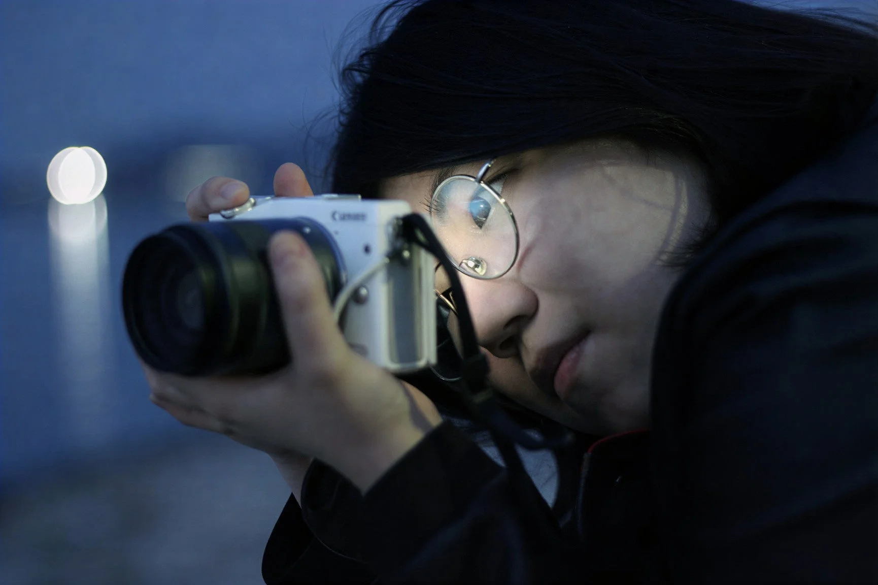 夜の屋外で、眼鏡をかけた女性が白いデジタルカメラを構え、熱心に何かを撮影している様子。