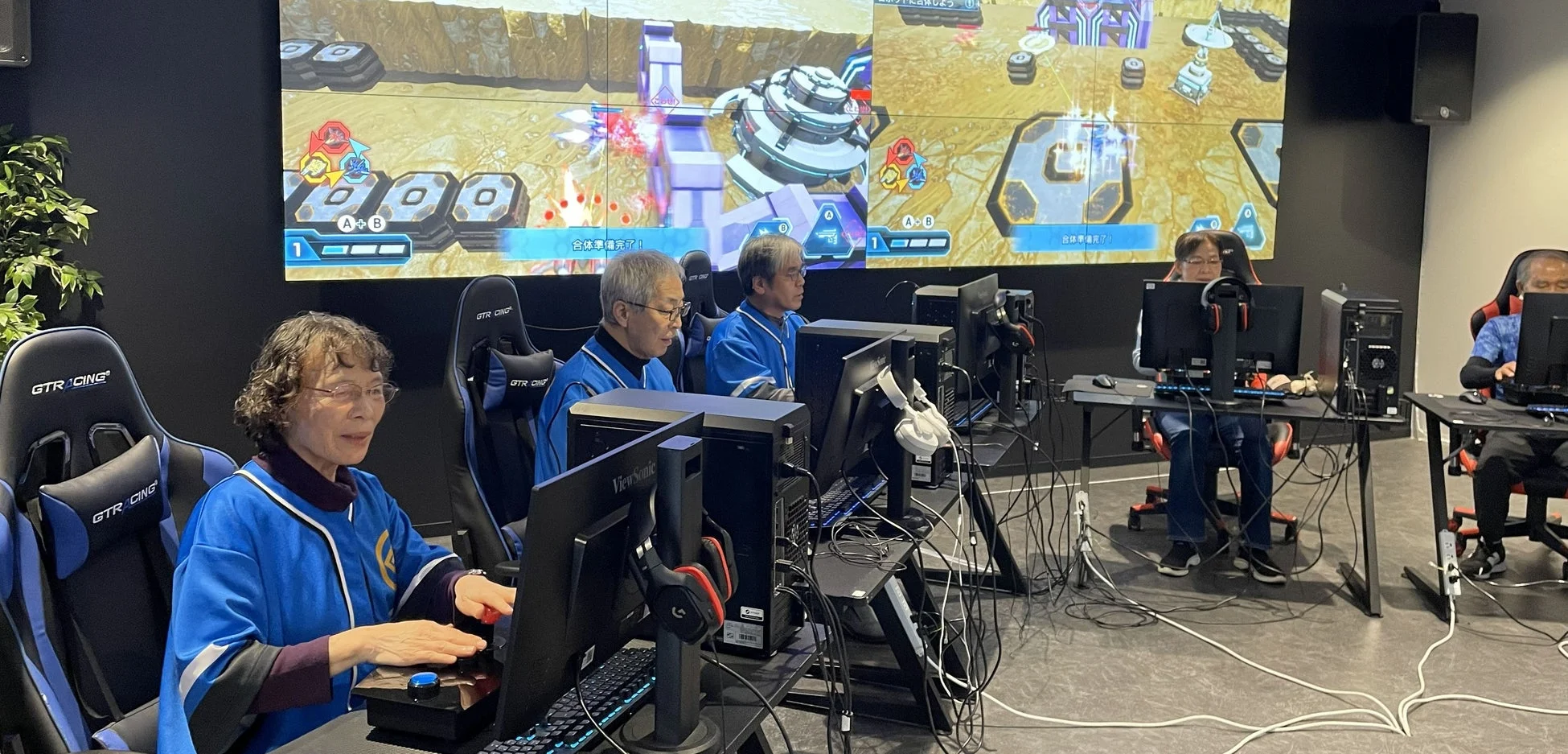 複数のシニア世代がゲーミングチェアに座り、PCゲームに熱中している様子
