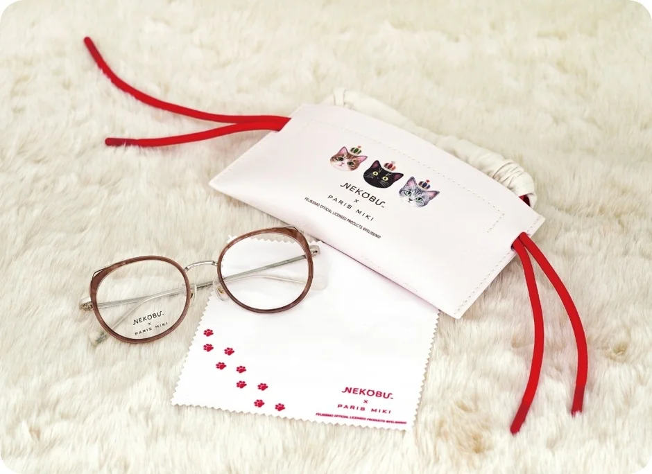 白いふわふわの背景に、猫のイラストが特徴的な眼鏡ケースと眼鏡拭き、そして茶色の縁の眼鏡が置かれています。ネコブとパリミキのコラボレーション商品で、可愛らしいデザインが目を引きます。
