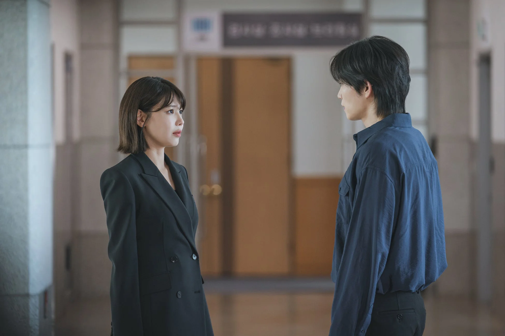 黒いブレザーを着た女性と青いシャツを着た男性が屋内で向かい合って立っており、真剣な表情で会話をしているか対峙しているような様子が捉えられています。