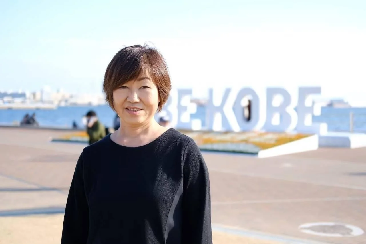 神戸のウォーターフロントにある「BE KOBE」モニュメントを背景に、笑顔で立つ女性のポートレートです。晴れた日の観光地の様子が伝わってきます。