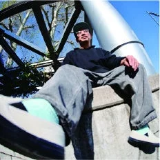 カジュアルな服装の男性がコンクリートの縁に座っている様子を、広角レンズで下から捉えた写真。青空の下、背景には金属製の構造物や木々が見える。