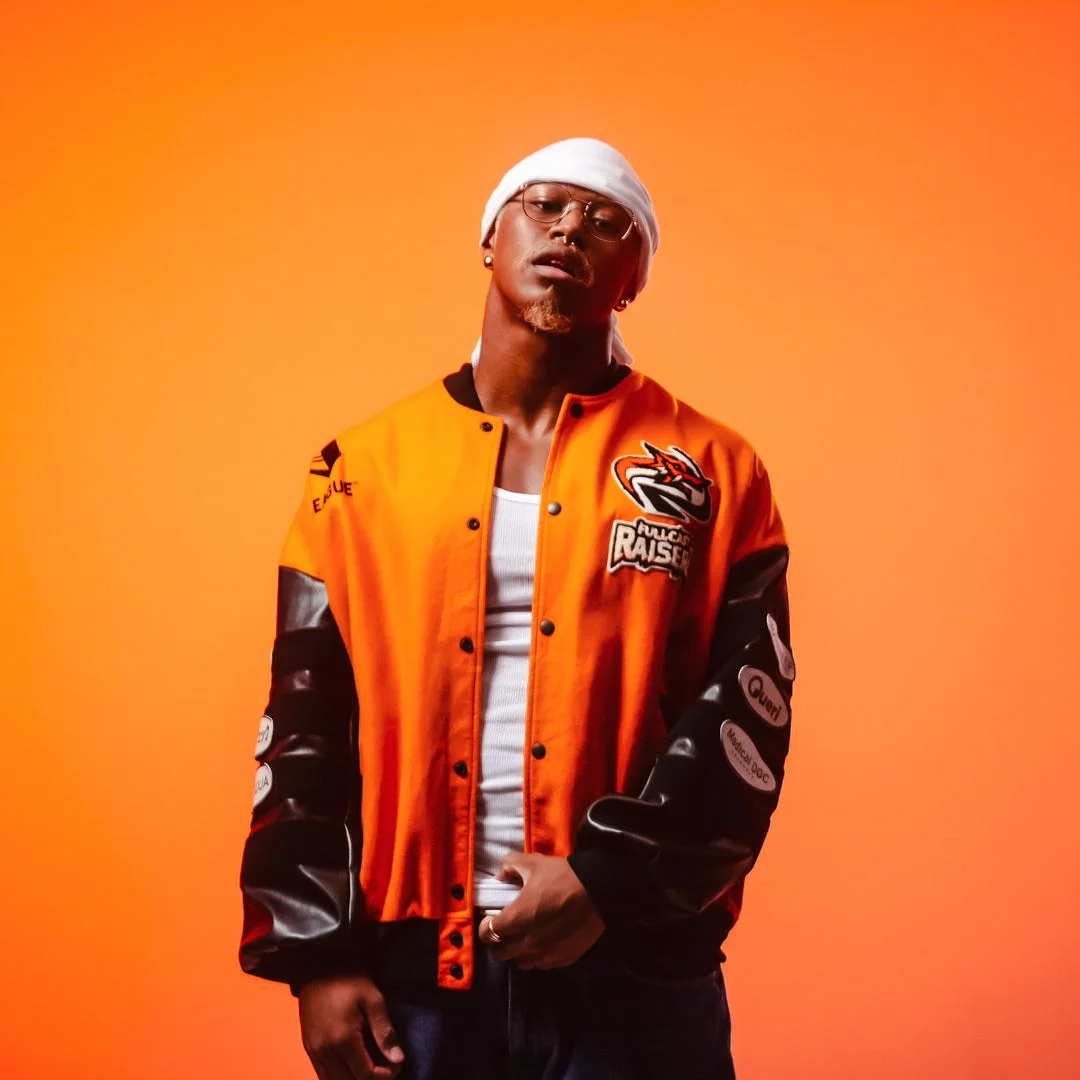 オレンジ色の背景をバックに、オレンジと黒のスタジャンと白いヘッドバンドを身につけた男性