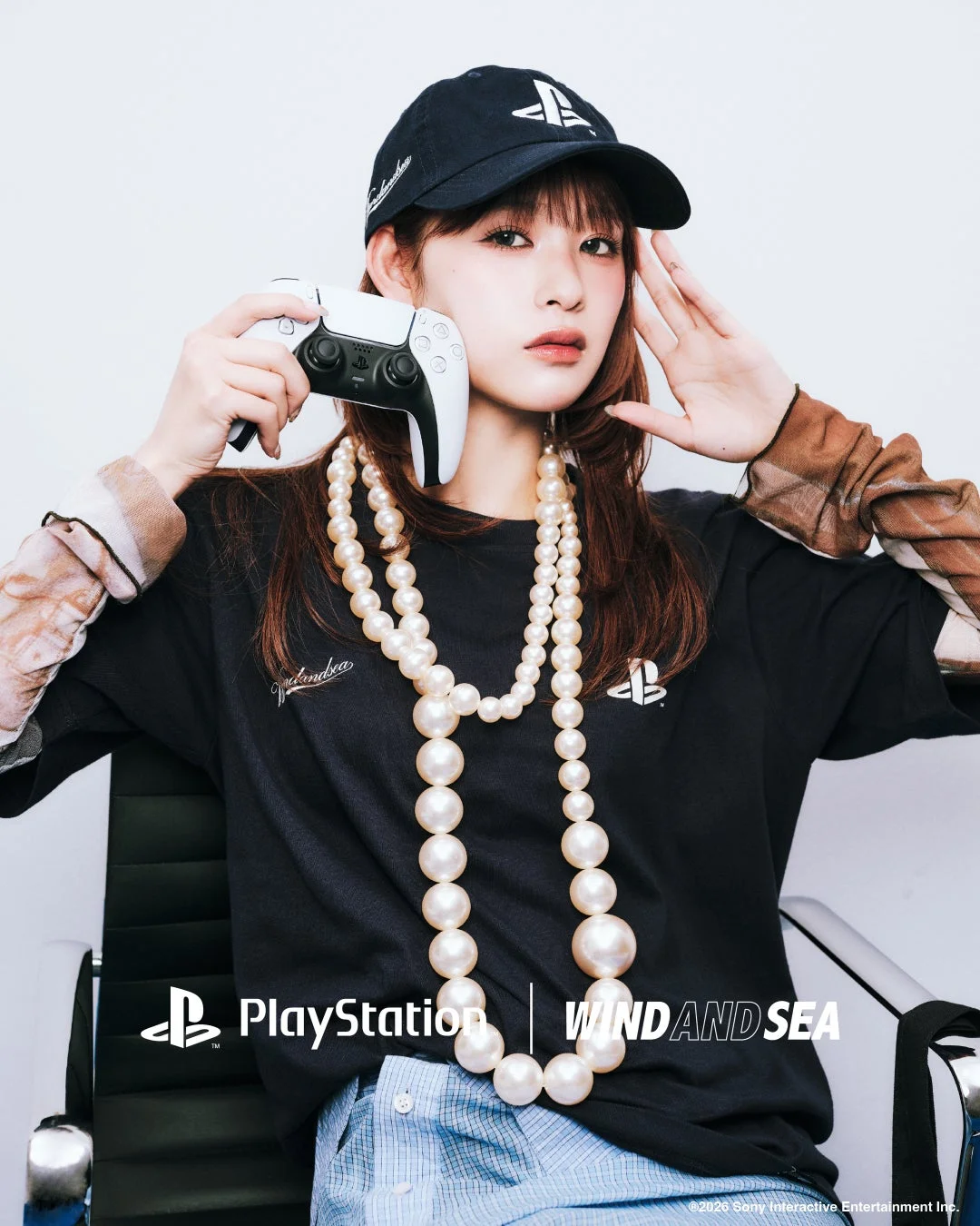PlayStationのキャップとTシャツを着用し、長いパールネックレスを身につけた若い女性が白いDualSenseコントローラーを持っている画像です。PlayStationとWIND AND SEAのコラボレーションをアピールしています。