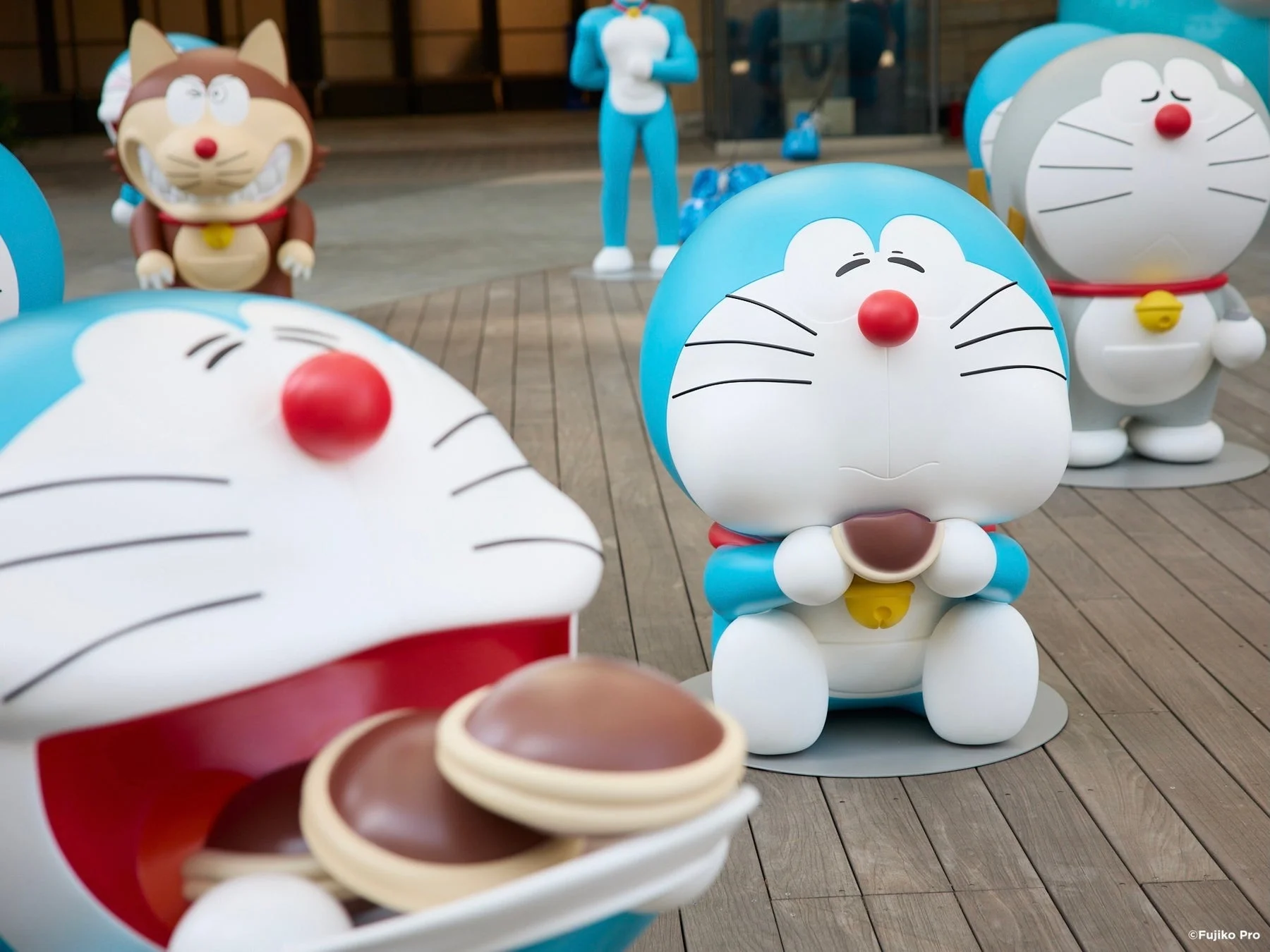 屋外の木製デッキに、ドラえもんの様々なキャラクターの像が展示されている様子
