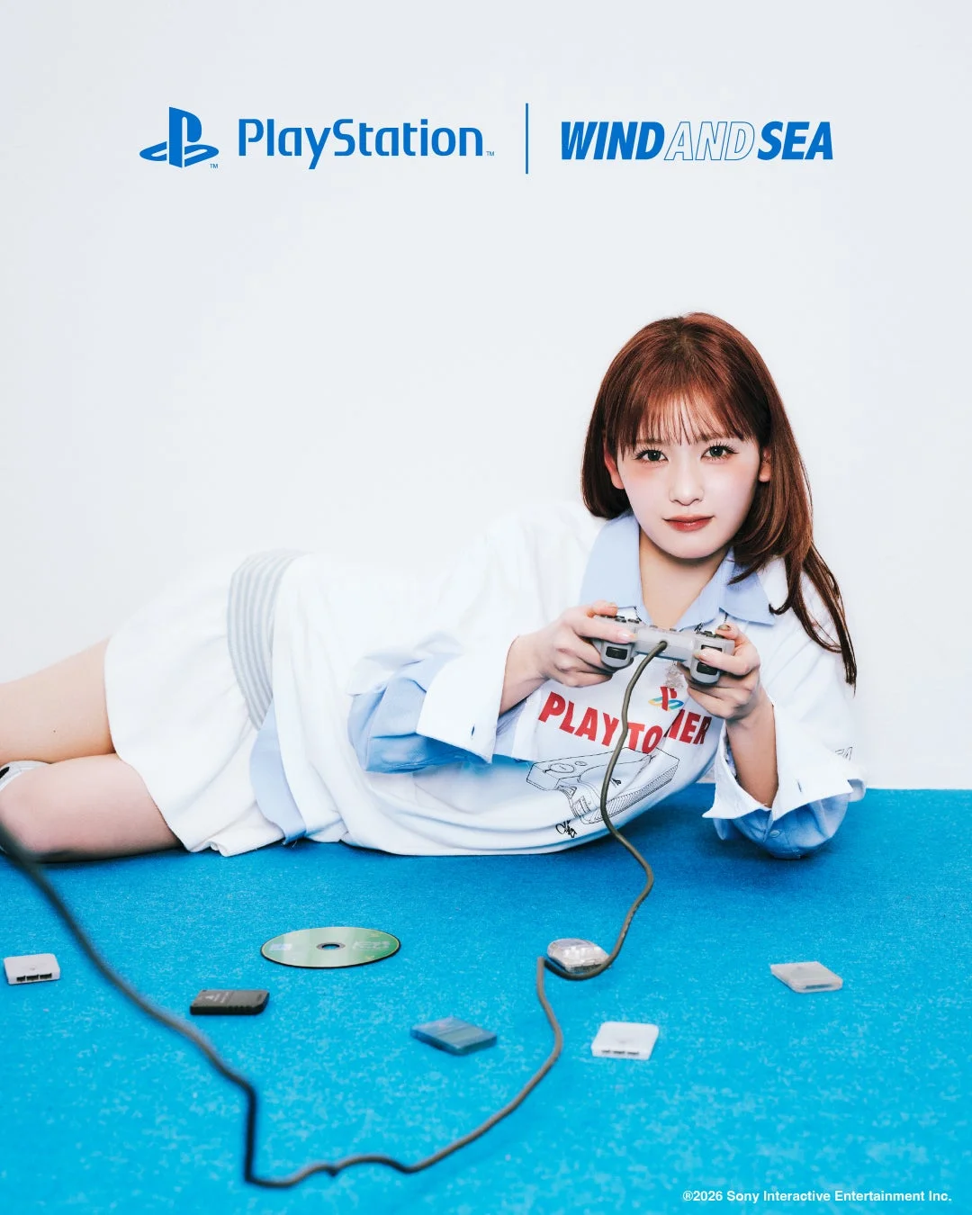 PlayStationとWIND AND SEAのコラボレーションを示す広告画像。青い床に横たわる女性がPlayStationのコントローラーを持ち、カメラを見つめています。周囲にはゲームディスクやメモリーカードが散らばっています。