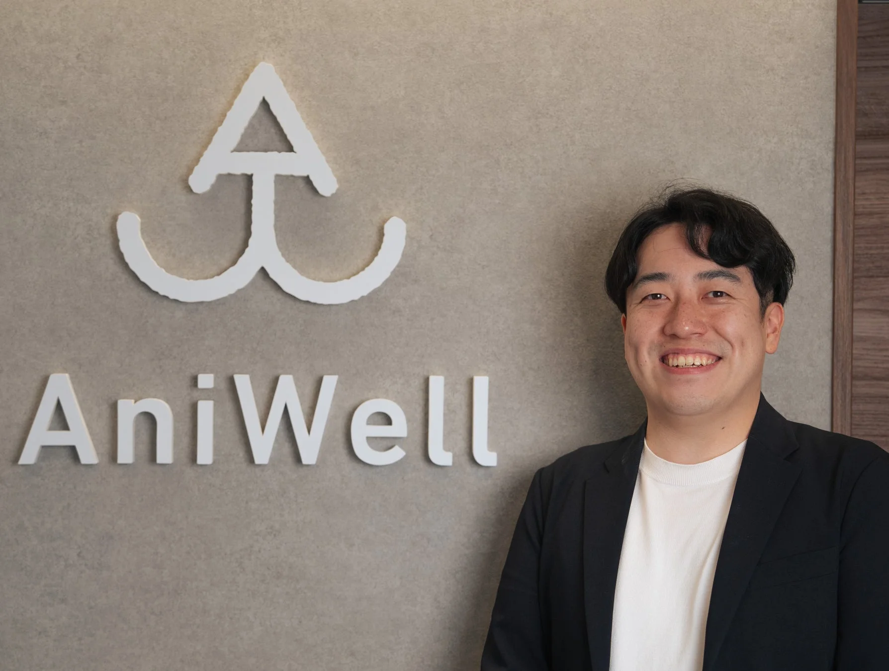 AniWellのロゴの前で笑顔で立つ男性