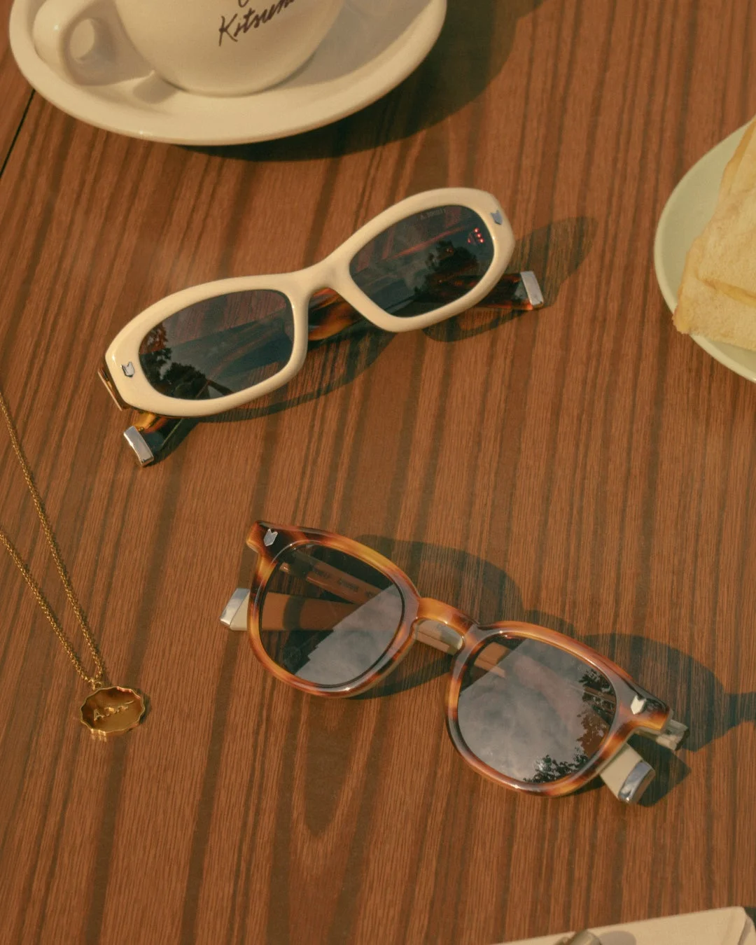 木目のテーブルに置かれたサングラスとべっ甲柄のサングラス、コーヒーカップ、トースト、ネックレス