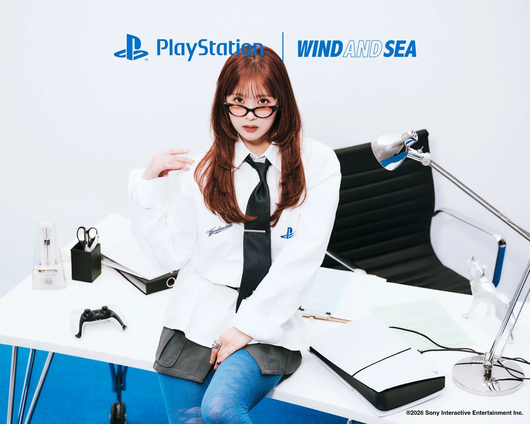 PlayStationとWIND AND SEAのコラボレーションをPRする画像。メガネをかけた女性が、オフィスのような空間でPS5とコントローラーと共にデスクに座っています。シャツには両ブランドのロゴが見えます。
