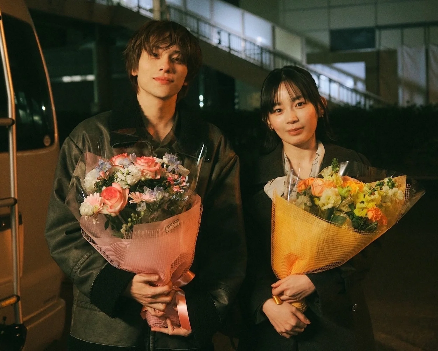 夜の屋外で、若い男女がそれぞれ美しい花束を抱えてカメラに微笑みかけています。温かく、お祝いのような雰囲気が感じられるポートレートです。