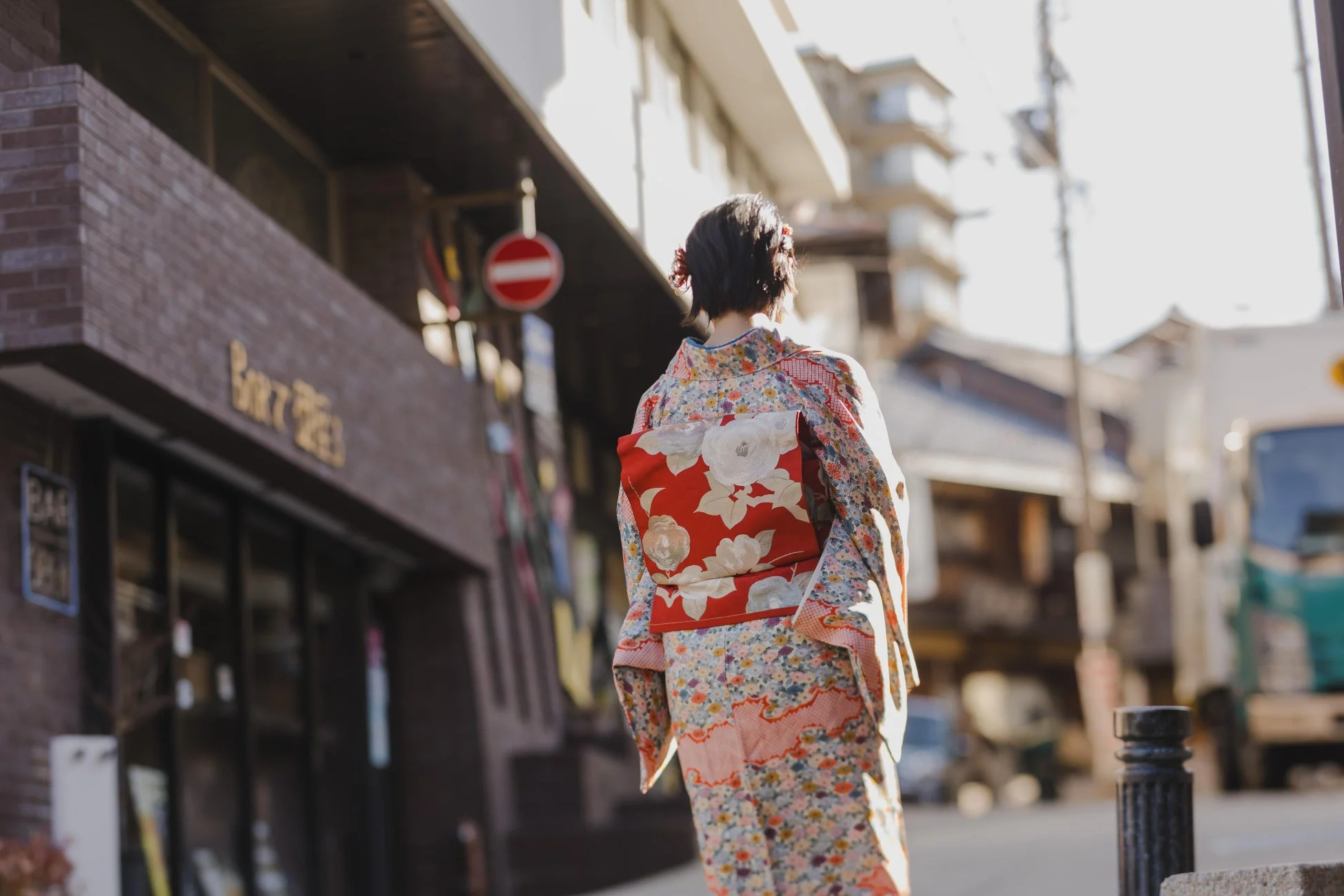美しい花柄の着物を着た女性が、日差しの当たる日本の街並みを歩いている後ろ姿です。赤い帯が印象的で、伝統的な風景に溶け込んでいる様子が伺えます。