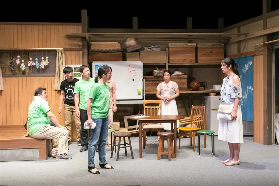 舞台上の和風のセットで、7人の登場人物が会話をしている演劇の一場面。木製の家具や棚が配置され、日常の風景を思わせる雰囲気です。