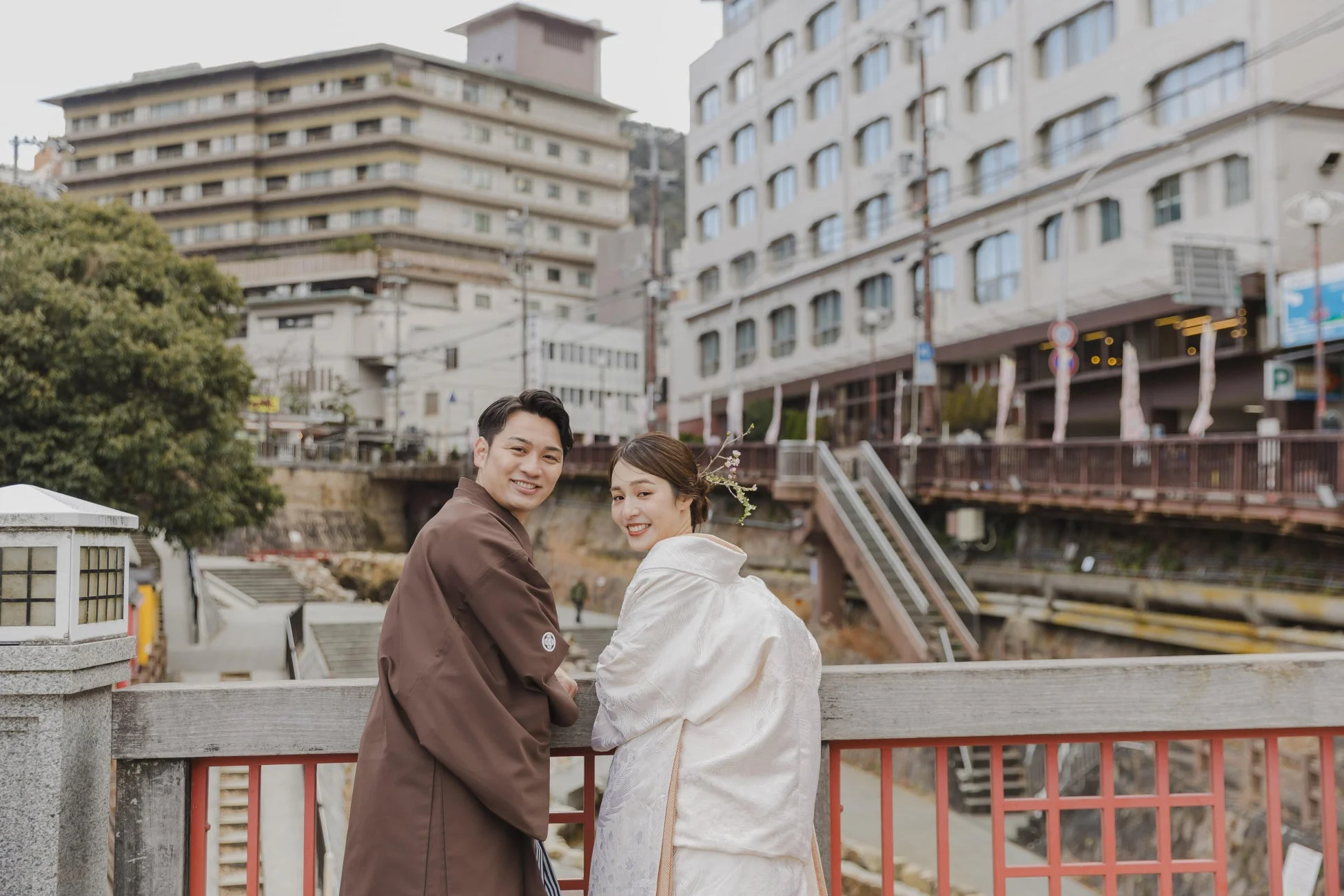 和装のカップルが笑顔でカメラを見つめているポートレート写真。背景には日本の街並みと建物が広がり、橋の上で撮影されたようです。