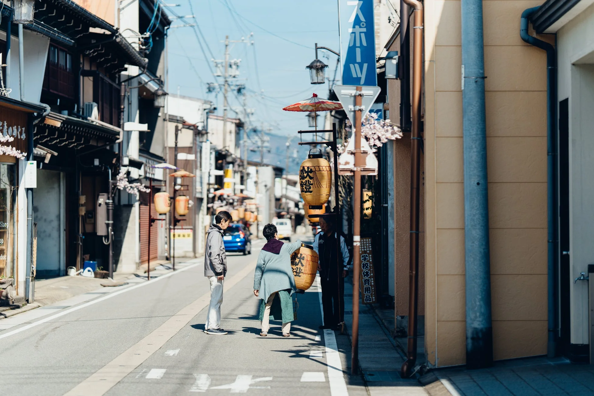 日本の伝統的な街並みを捉えた写真。青空の下、商店が並ぶ通りを数人の人々が歩いている。通りには「スポーツ」の看板や「大安吉日」と書かれた提灯が吊るされ、桜の花も彩りを添えている。右側には「学校体育用品」の看板が見える。