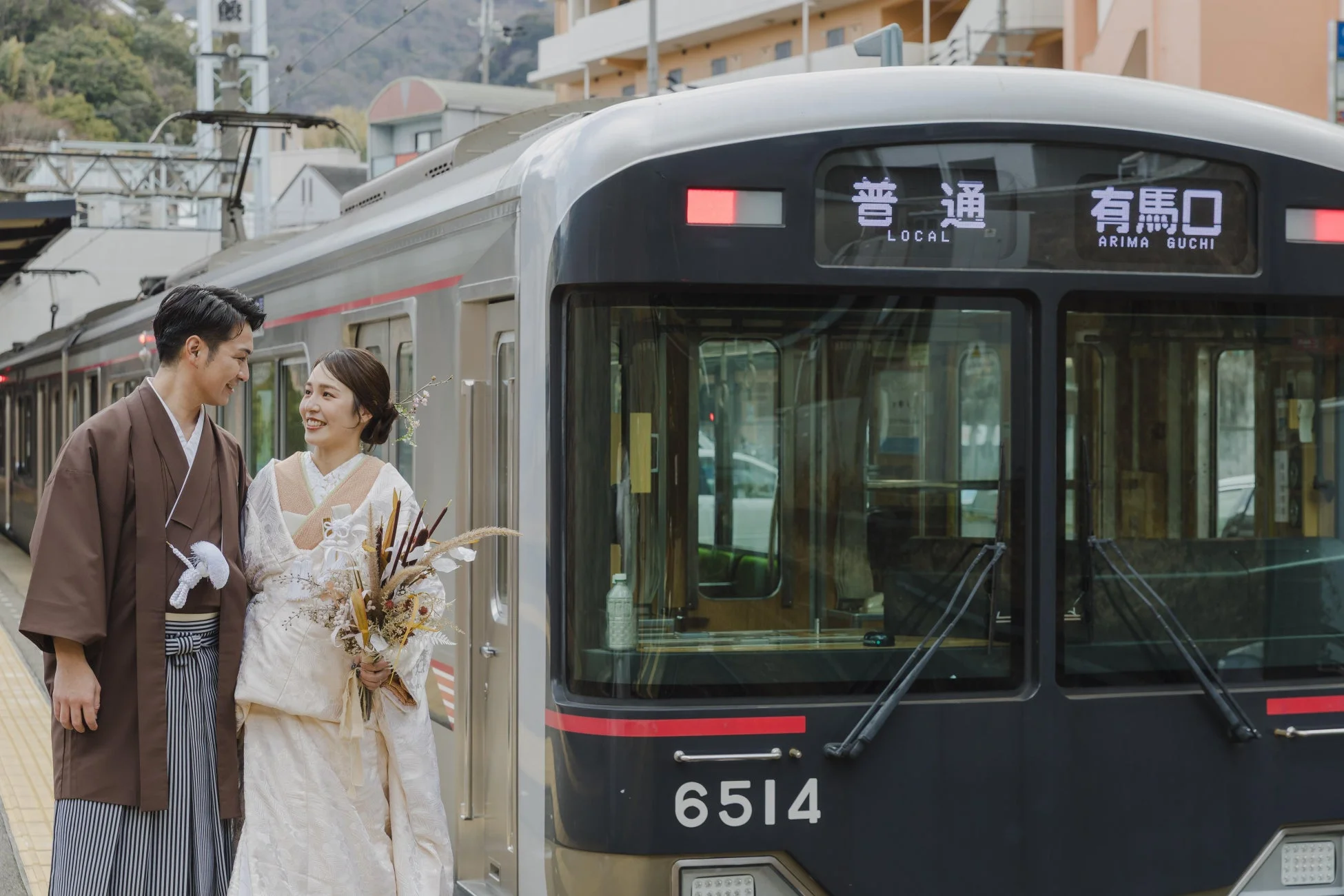 駅のホームで和装のカップルが笑顔で向かい合っている。女性は白い着物にブーケを持ち、男性は茶色の着物と袴を着用。背景には「普通 有馬口」と表示された電車が停車しており、結婚式の前撮りのようなロマンチックな雰囲気が漂う。