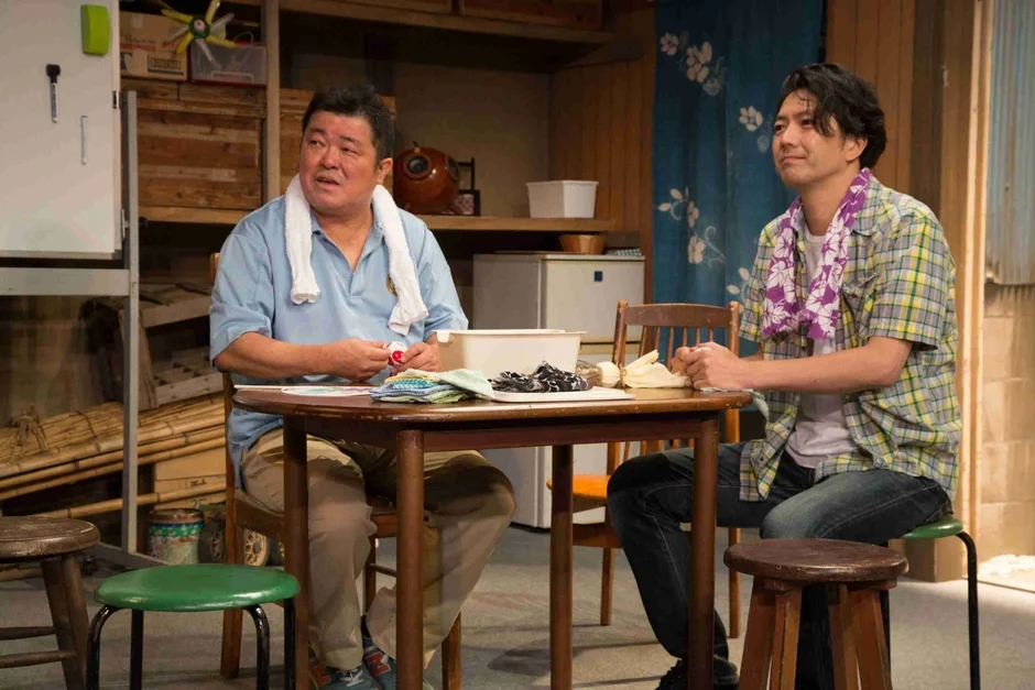 二人の男性が木製のテーブルを囲んで座っており、何か作業をしているか、会話を楽しんでいる様子です。一人は水色のポロシャツに白いタオル、もう一人はチェックのシャツに紫色の柄物タオルを首にかけており、リラックスした雰囲気が伝わります。