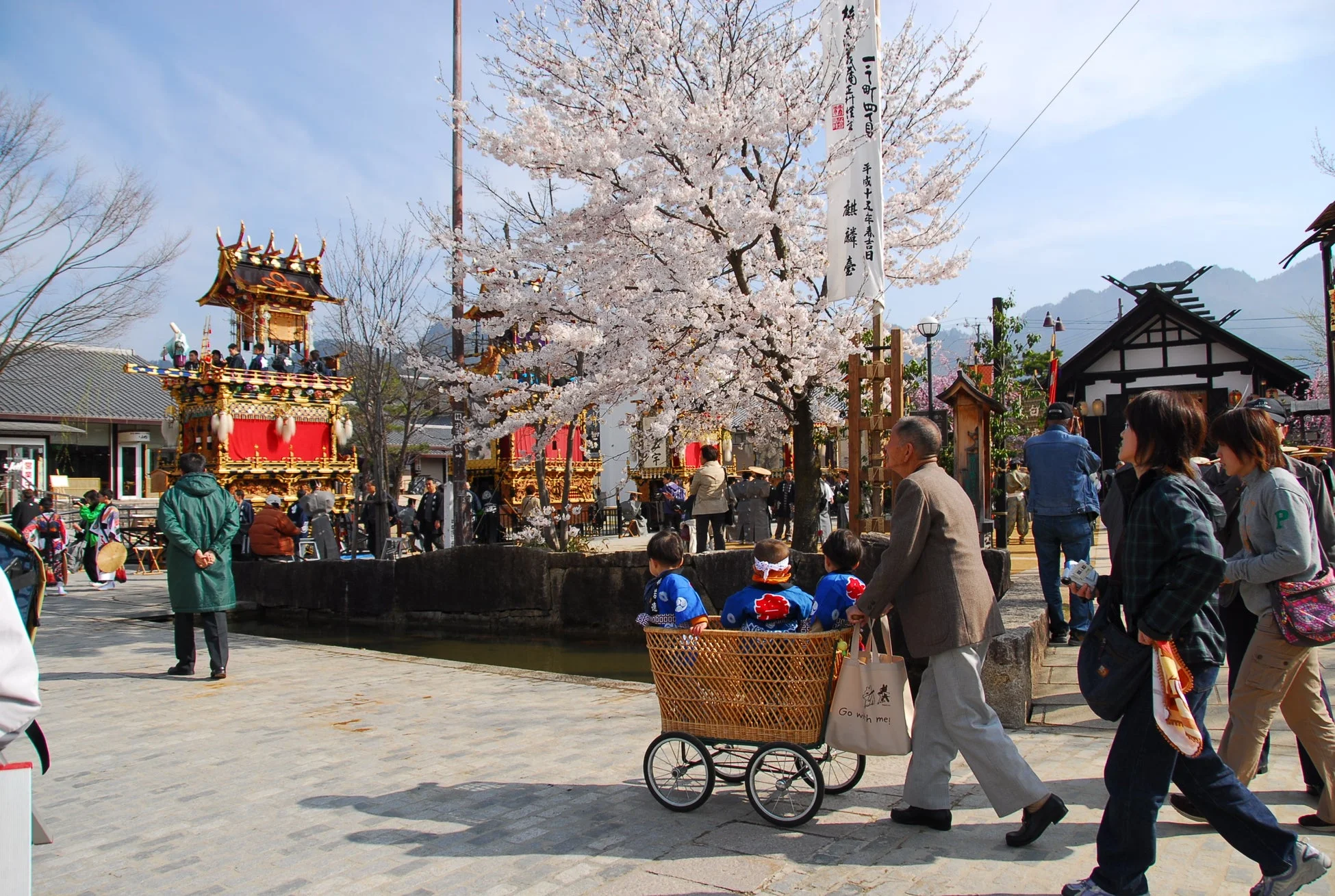 満開の桜が咲き誇る日本の春の祭りの情景。豪華な山車が並び、多くの人々で賑わう中、子供たちを乗せた手押し車を押す男性が通りを歩く、活気ある一日が描かれている。