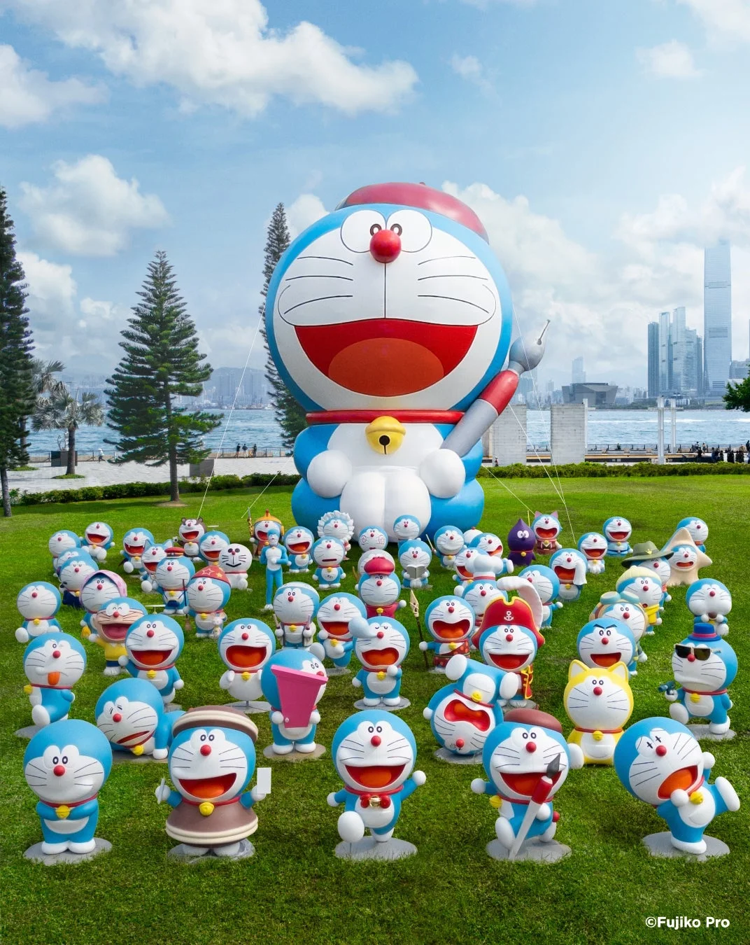 広々とした芝生の公園で、巨大なドラえもんの像を中心に、様々な衣装を着た多数の小さなドラえもんの像が展示されている様子
