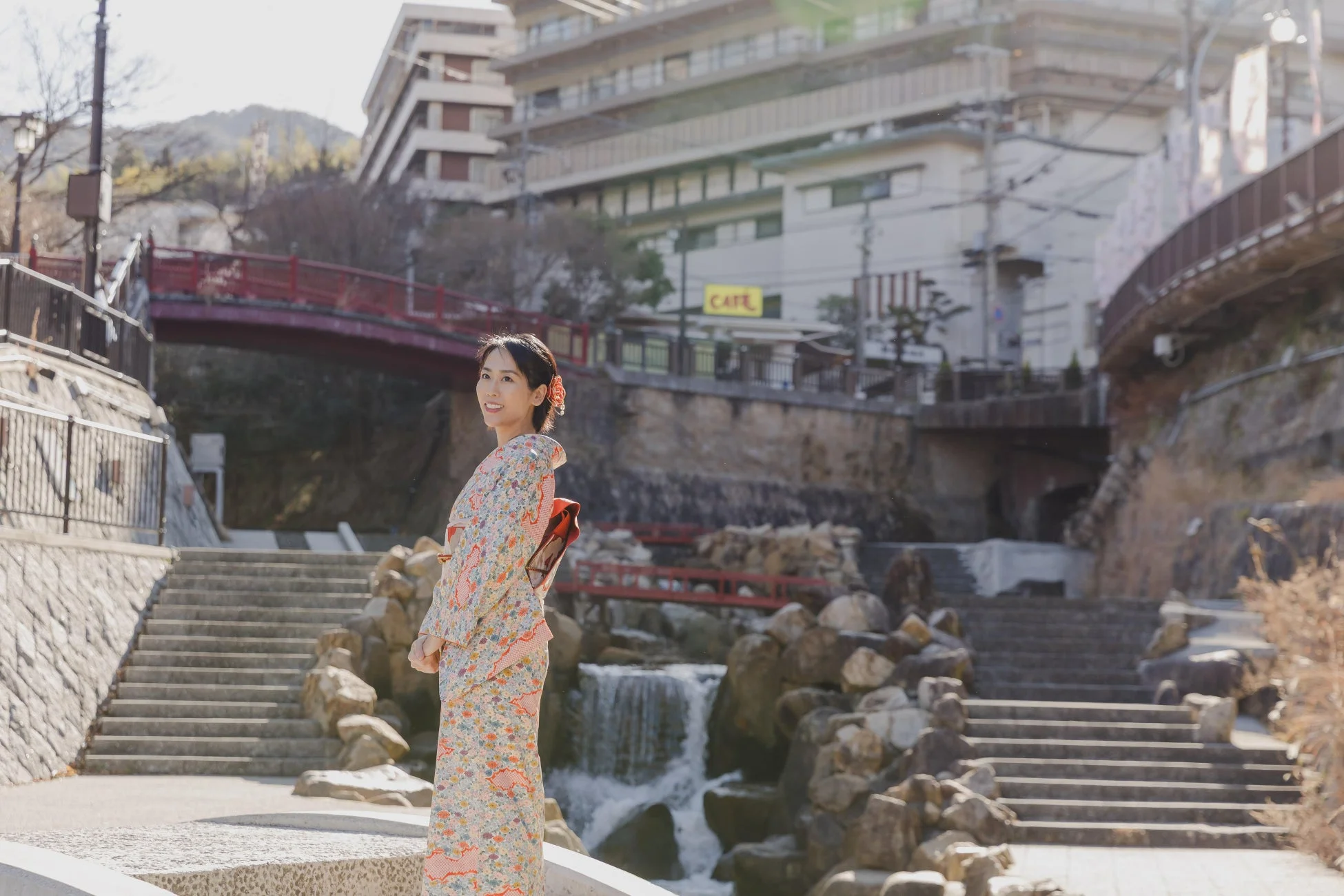 着物姿の女性が笑顔で振り返る、日本の風情ある風景です。背景には滝や階段、赤い橋が見え、和やかな観光地の雰囲気が伝わってきます。