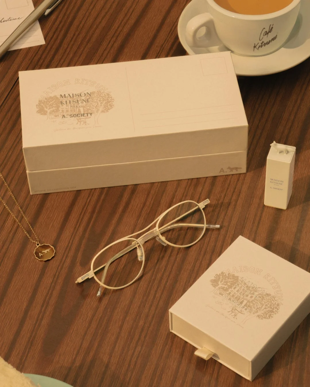Maison KitsunéとA. Societyのロゴが入った白い箱と小さなパッケージ、メガネ、ネックレスが木目調のテーブルに置かれている