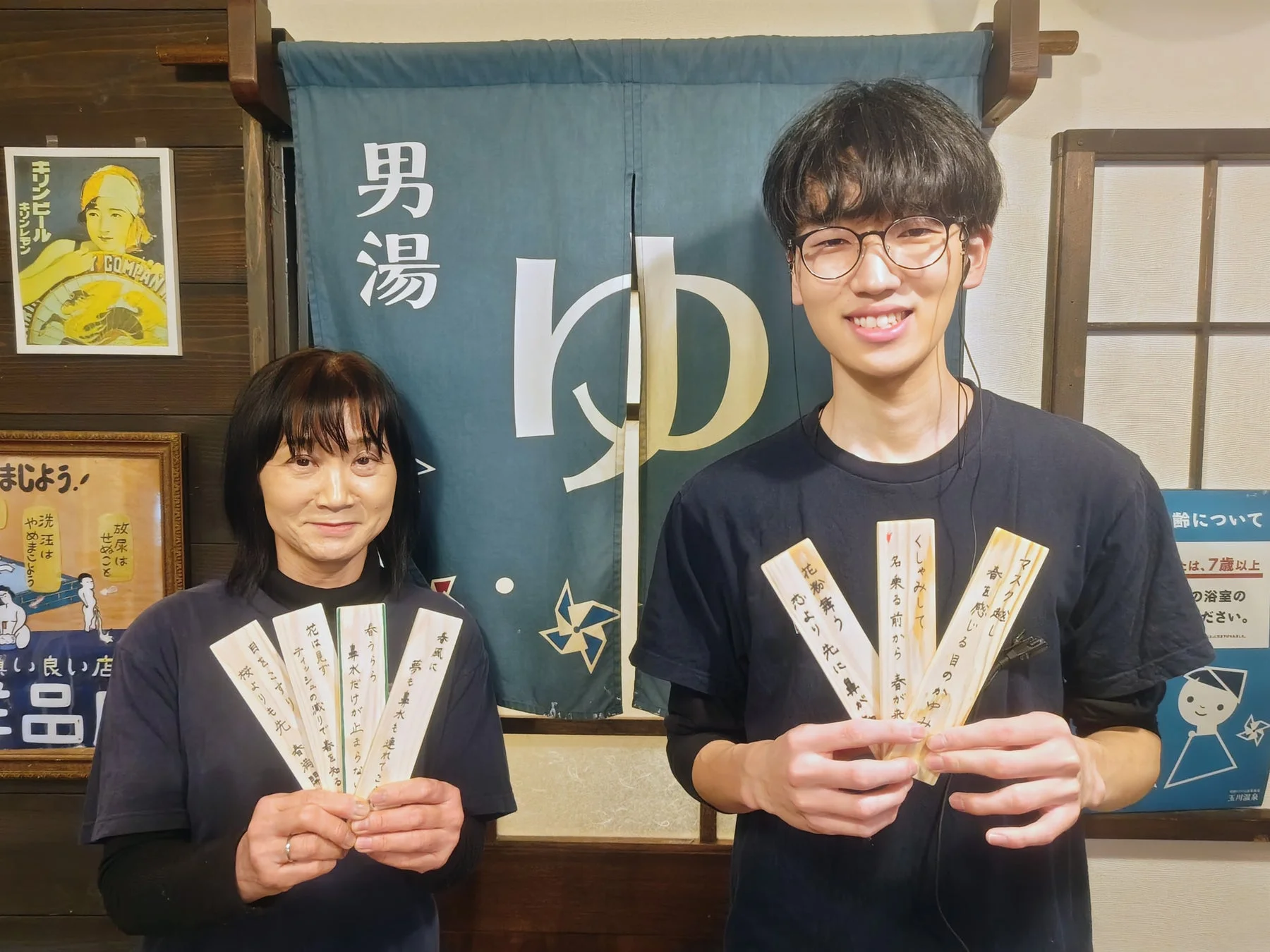 男湯ののれんの前で川柳の木札を持つ男女