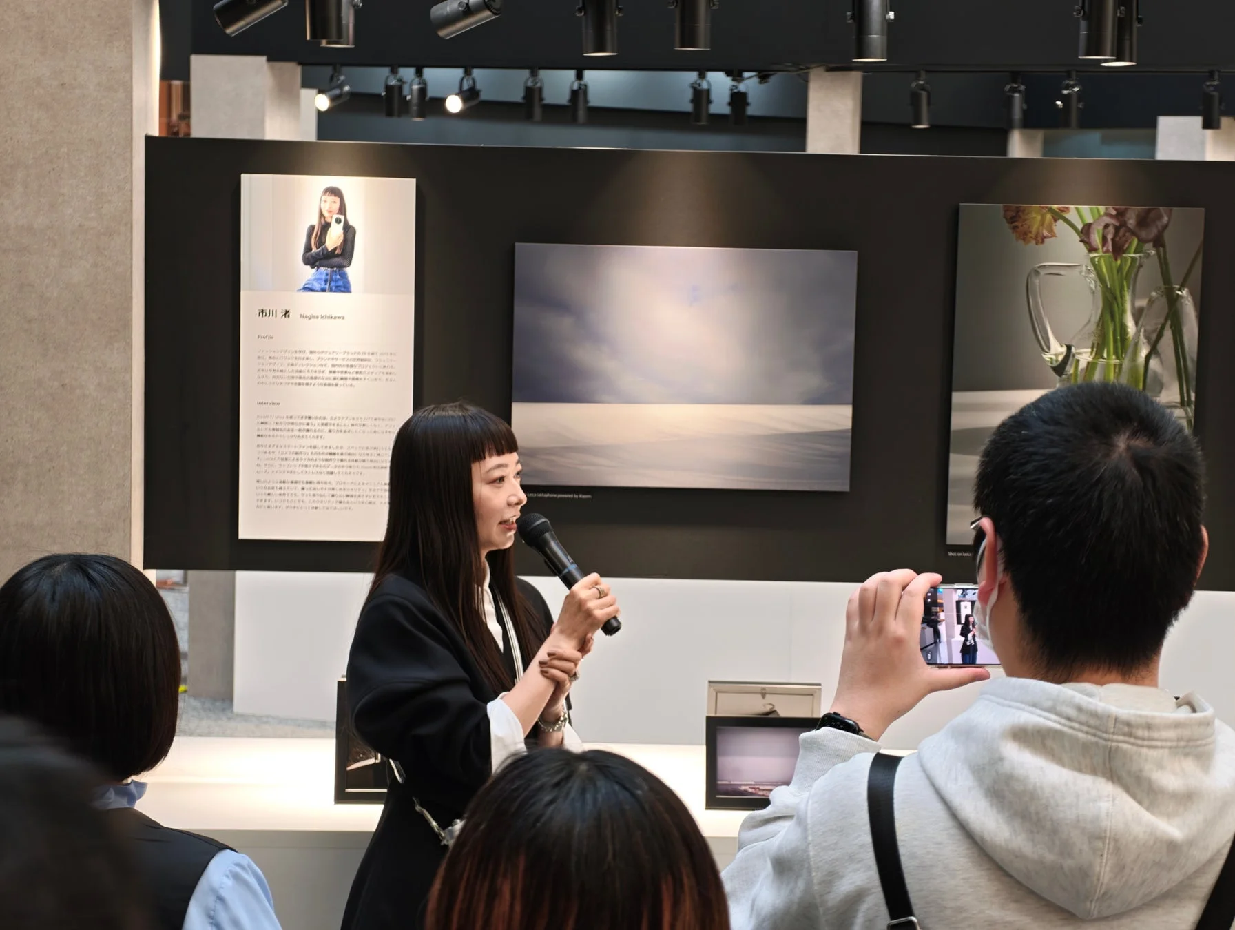 市川渚氏が写真展または製品発表会で講演しており、聴衆が熱心に耳を傾けたりスマートフォンで撮影したりしている様子