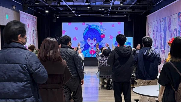 大画面に映し出された青い髪のアニメキャラクターを多くの観客が見つめています。一部の観客は手を挙げており、キャラクターとの交流やイベントへの参加を楽しんでいる様子がうかがえます。