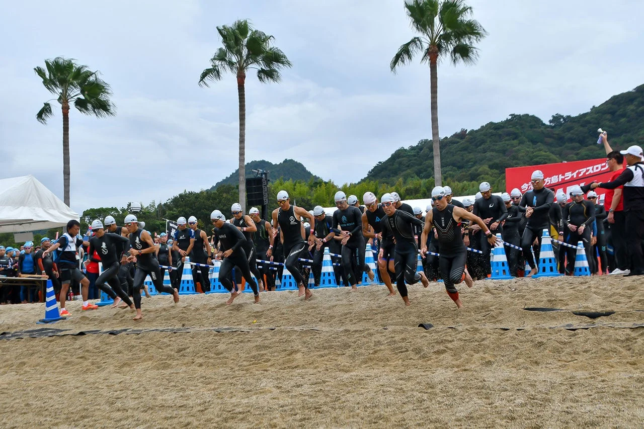 砂浜でトライアスロン競技がスタートし、ウェットスーツを着た多くの選手が一斉に走り出している様子