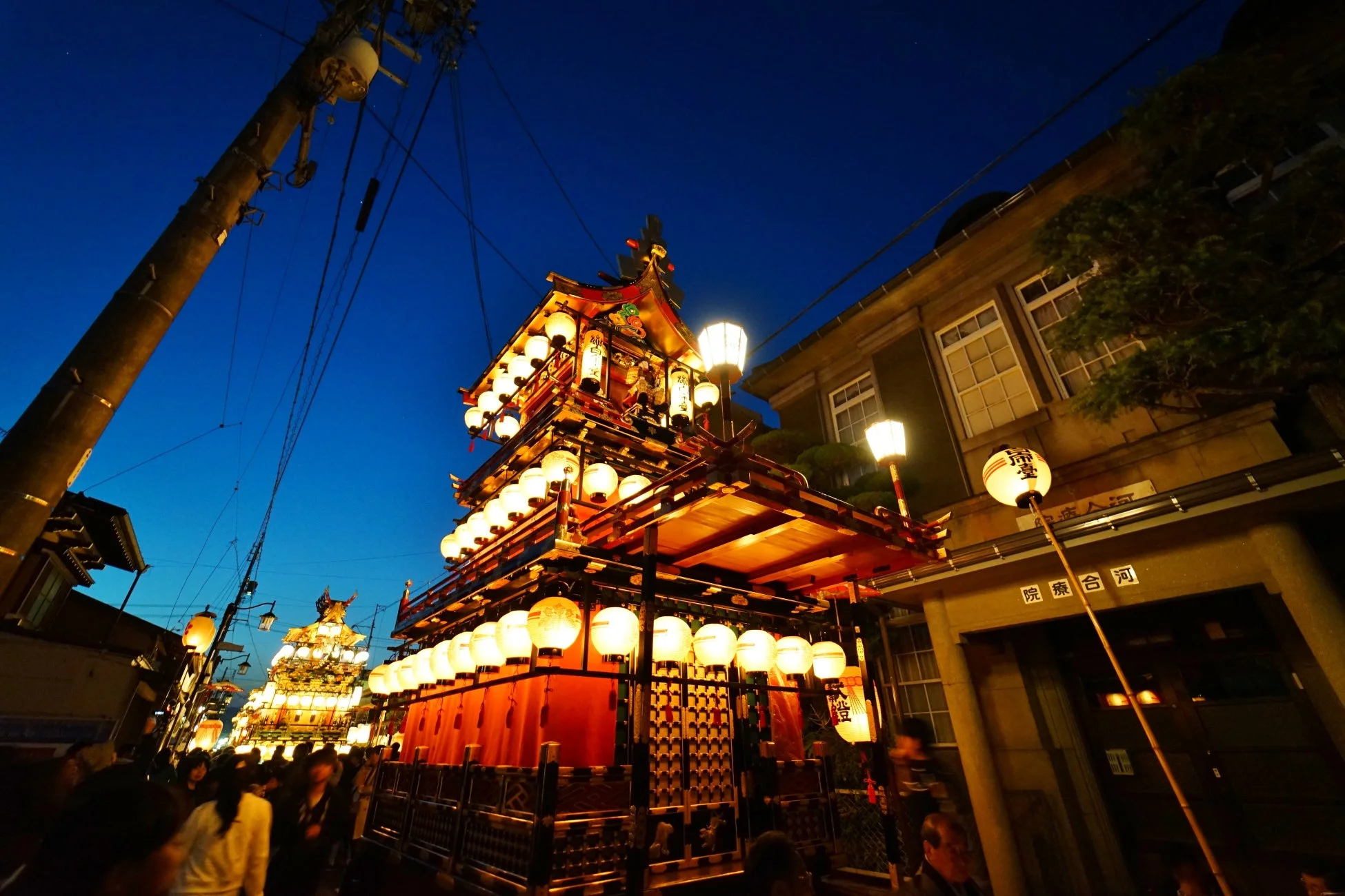 夜空の下、提灯で煌びやかに装飾された山車が通りを練り歩く日本の伝統的な祭りの様子。多くの人々が集い、幻想的な雰囲気に包まれている。